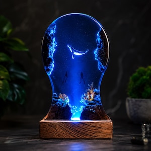 May include: A teardrop-shaped resin lamp featuring a deep blue underwater scene with a swimming whale, two scuba divers, and coral formations. The piece is illuminated from below and mounted on a rectangular wooden base with a natural grain finish.