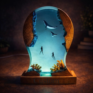 Op de afbeelding: Een handgemaakte lamp met een onderwaterlandschap. De lamp toont walvissen, duikers en koraal in heldere hars, omlijst door houten randen. De basis is van hout, met het woord "Custom" zichtbaar.