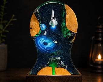 May include: A decorative lamp featuring a space scene encased in clear resin and natural wood. The design includes a white rocket ship, a small astronaut, a blue planet, and a swirling black hole set against a dark, starry background.