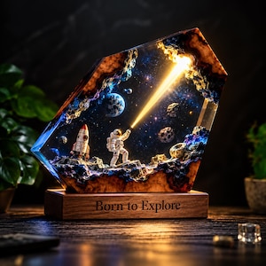 May include: A resin lamp featuring a space scene with an astronaut holding a light beam, a rocket, and planets. The piece is set in a geometric wood-encased frame with the text Born to Explore engraved on the rectangular wooden base.