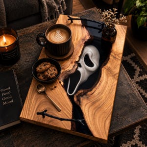May include: A rectangular wooden serving tray featuring a central river of black resin embedded with a white ghost-like mask. The tray includes two black branch-shaped metal handles, a dark ribbed mug with latte art, a small bowl of cookies, and a spoon.