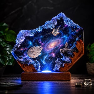 May include: A hexagonal resin and wood lamp featuring a deep space scene with a spiral galaxy, a miniature spaceship, and an astronaut standing on a rocky ledge. The piece is illuminated from below, casting a blue glow through the crystalline edges.