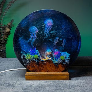 May include: A round resin lamp featuring a deep blue galaxy design with jellyfish and coral. The lamp sits on a wooden base and has a white power cord. The jellyfish are illuminated with blue and purple hues.