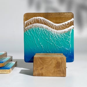 May include: Square wooden art piece depicting a beach scene. The top is wood-toned, transitioning to turquoise and blue resin, mimicking ocean waves. Several matching coasters are stacked in the foreground.