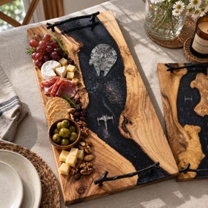 May include: A rectangular wooden serving tray with a central black resin river inlay featuring a galaxy pattern, a Millennium Falcon, and an X-wing starfighter. The tray has two black metal handles shaped like tree branches at the ends.