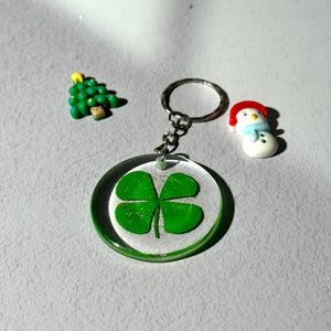 May include: A clear, round keychain with a green four-leaf clover design. Accompanying the keychain are small decorative figures of a green Christmas tree and a snowman. The items are set against a light gray background.