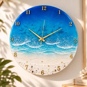 May include: A round wall clock featuring a resin ocean scene with blue water, white sea foam waves, and a sandy beach bottom. The clock face includes gold-colored numbers from 1 to 12, gold hands, and small gold star accents scattered on the sand.