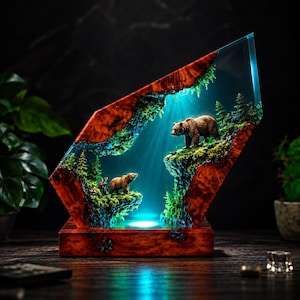Może przedstawiać: Dekoracyjna lampa wykonana z polerowanego czerwonobrązowego drewna i przezroczystej żywicy, przedstawiająca miniaturową leśną scenę z dwoma brązowymi niedźwiedziami na skalnych półkach. Podstawa zawiera źródło światła oświetlające wnętrze na niebiesko.