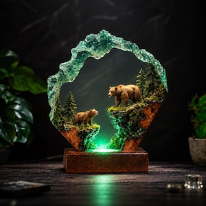 Może przedstawiać: Ozdobna lampa z dwiema figurkami niedźwiedzi brunatnych stojącymi na skalistych, porośniętych mchem klifach z miniaturowymi sosnami. Scena jest otoczona półprzezroczystym, teksturowanym turkusowym łukiem z żywicy, osadzonym na prostokątnej drewnianej podstawie.