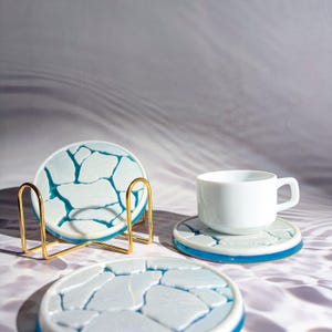 May include: A set of round coasters with a white and teal cracked ice design, a white teacup, and a gold metal coaster holder. The coasters have a blue border. The teacup sits on a matching coaster.