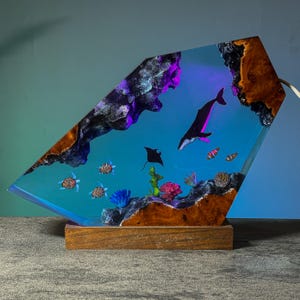 May include: A decorative lamp featuring an underwater scene with a whale, stingray, and sea turtles. The lamp has a wooden base and a clear resin body with colorful coral and rock formations. The lamp is lit with blue and purple lights.