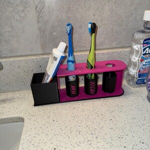 May include: A pink and black toothbrush holder with two toothbrushes and a tube of toothpaste. The holder has slots for toothbrushes and a small container for toothpaste. The holder has the names "PENN" and "POTTER" on the front.