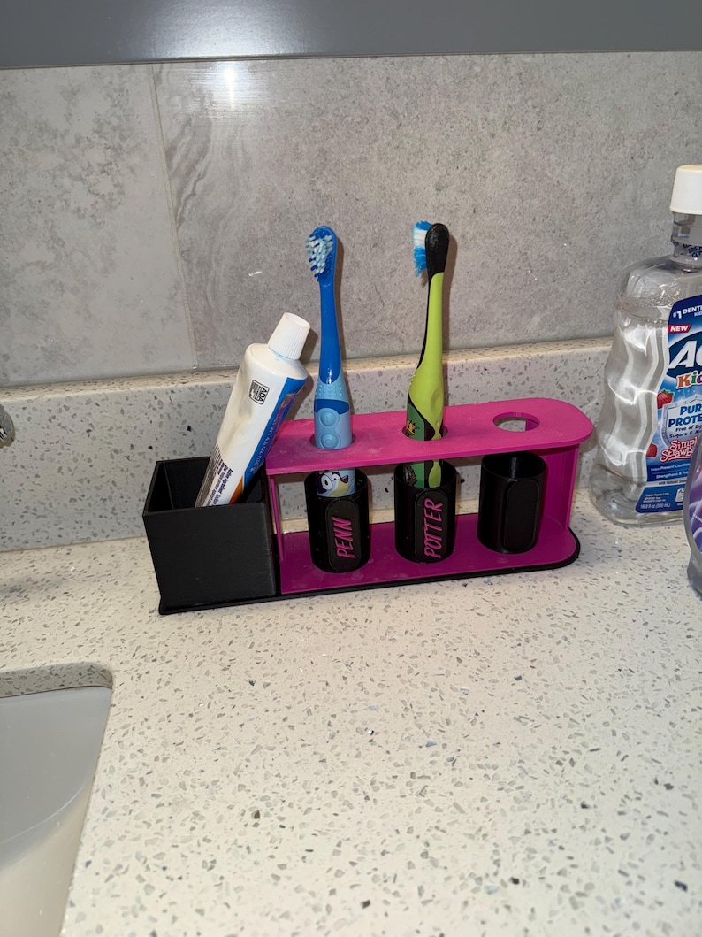 May include: A pink and black toothbrush holder with two toothbrushes and a tube of toothpaste. The holder has slots for toothbrushes and a small container for toothpaste. The holder has the names "PENN" and "POTTER" on the front.