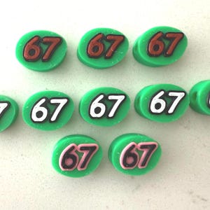 May include: Ten green, oval-shaped beads with the number 67 in various colors. Some have white numbers with a black outline, others have brown or pink numbers with a black outline. The beads are arranged on a white surface.
