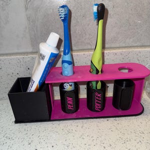 May include: A pink and black toothbrush holder with two toothbrushes and a tube of toothpaste. The holder has slots for toothbrushes and small cups labeled "PENN" and "POTTER".