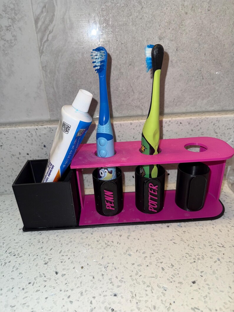 May include: A pink and black toothbrush holder with two toothbrushes and a tube of toothpaste. The holder has slots for toothbrushes and small cups labeled "PENN" and "POTTER".