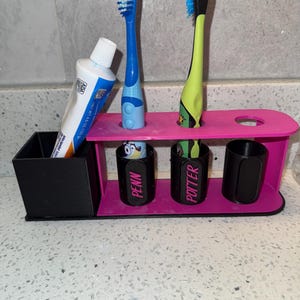 May include: A pink and black toothbrush holder with two toothbrushes and a tube of toothpaste. The holder has slots for toothbrushes and cups labeled "PENN" and "POTTER".