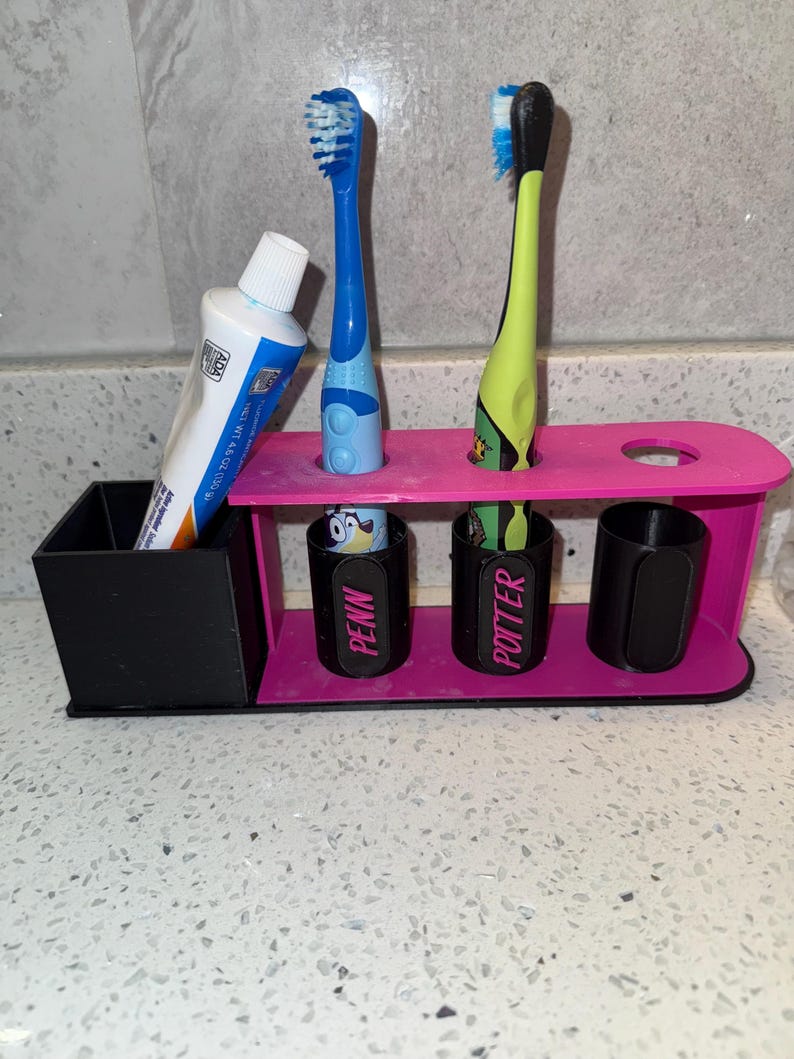 May include: A pink and black toothbrush holder with two toothbrushes and a tube of toothpaste. The holder has slots for toothbrushes and cups labeled "PENN" and "POTTER".