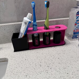 May include: A pink and black toothbrush holder with space for toothbrushes, toothpaste, and cups. The holder has two blue toothbrushes and one green toothbrush. The cups are labeled "PENN" and "POTTER".