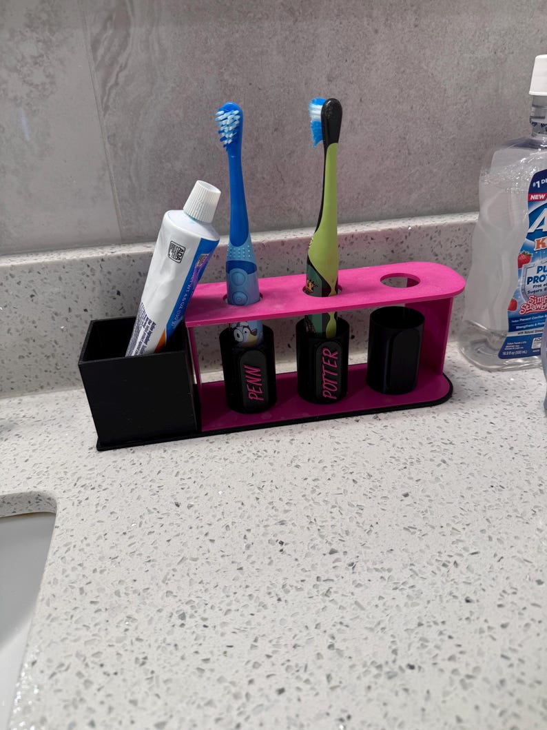 May include: A pink and black toothbrush holder with space for toothbrushes, toothpaste, and cups. The holder has two blue toothbrushes and one green toothbrush. The cups are labeled "PENN" and "POTTER".