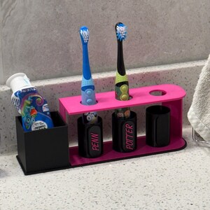 May include: A pink toothbrush holder with two toothbrushes and a black container for toothpaste. The toothbrushes are blue and black with colorful bristles. The holder has the names "PENN" and "POTTER" on the toothbrush holders.