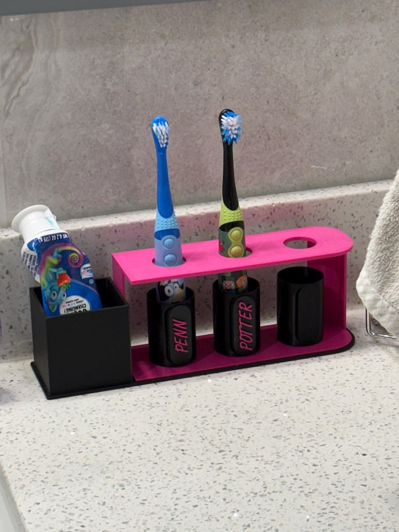May include: A pink toothbrush holder with two toothbrushes and a black container for toothpaste. The toothbrushes are blue and black with colorful bristles. The holder has the names "PENN" and "POTTER" on the toothbrush holders.