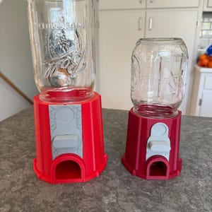 May include: Two candy dispensers with bright red plastic bases and clear glass Ball mason jars attached to the top. Each dispenser features a light gray plastic dispensing mechanism with a lever and a small opening at the bottom for items to exit.