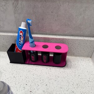 May include: A pink and black toothbrush holder with a tube of Crest Kids toothpaste and a blue toothbrush. The holder has three slots for toothbrushes and three black containers with the names "PENN" and "POTTER" on them.