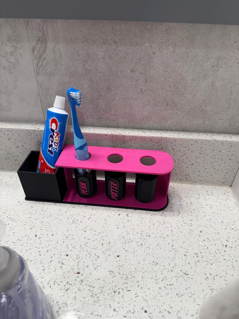 May include: A pink and black toothbrush holder with a tube of Crest Kids toothpaste and a blue toothbrush. The holder has three slots for toothbrushes and three black containers with the names "PENN" and "POTTER" on them.