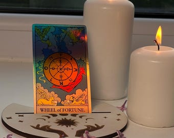 May include: A tarot card, featuring the text "Wheel of Fortune", rests on a wooden stand, with two lit white candles. The card has colourful illustrations. Lilac flowers are scattered around the base.