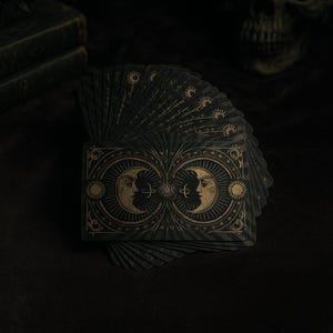 Può includere: Un mazzo di tarocchi con un design verde scuro e oro. Le carte presentano illustrazioni di lune, soli e simboli celesti. Le carte sono a ventaglio, rivelando gli intricati dettagli dell'opera d'arte. L'estetica generale è gotica e mistica.