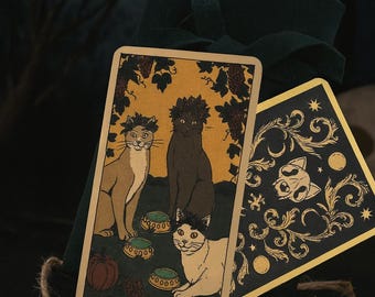 May include: Tarot card featuring three cats, with the text "THREE OF CUPS". The card has a beige background with a dark green velvet bag in the background. A second card with a cat skull design is also visible.