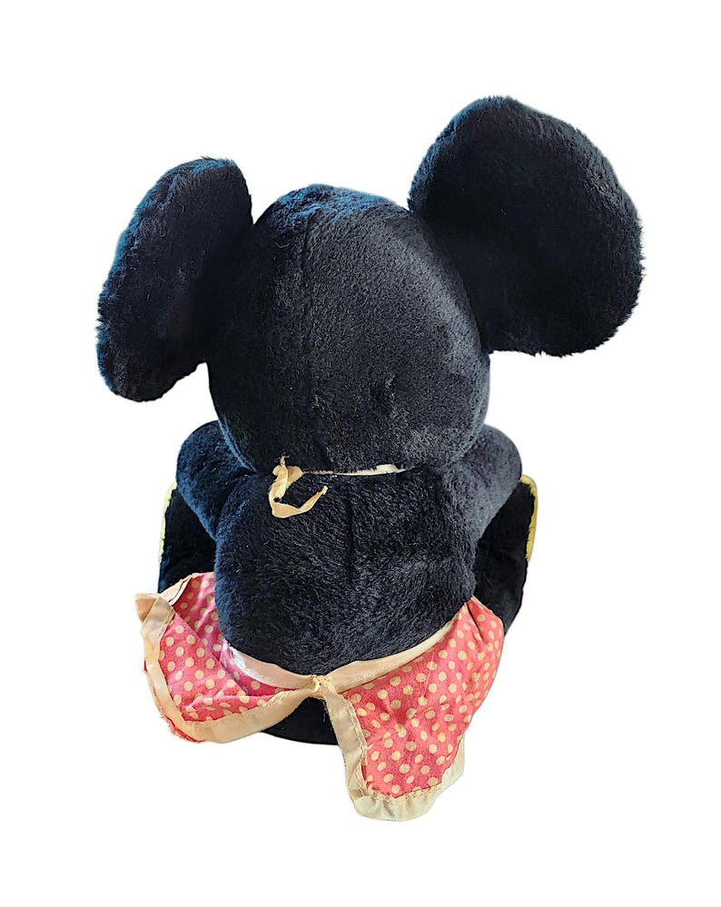 May include: A vintage black plush toy mouse with large, rounded ears. The mouse wears a pink and white polka-dot skirt with a cream-colored trim. The toy is viewed from the back, showcasing its plush texture and design.