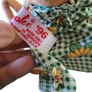 Może przedstawiać: Zbliżenie na metkę lalki z napisem "alee" i rokiem "'96" w kolorze czerwonym. Na metce widnieje napis "MADE ONLY IN MEREDITH, NEW HAMPSHIRE, U.S.A." Sukienka lalki jest zielono-biała z kwiatowym wzorem.