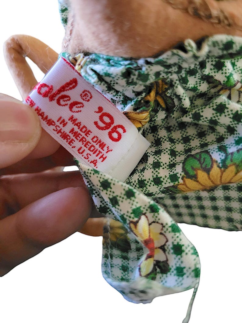 Może przedstawiać: Zbliżenie na metkę lalki z napisem "alee" i rokiem "'96" w kolorze czerwonym. Na metce widnieje napis "MADE ONLY IN MEREDITH, NEW HAMPSHIRE, U.S.A." Sukienka lalki jest zielono-biała z kwiatowym wzorem.