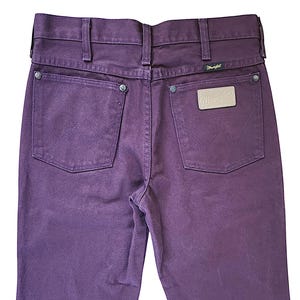 Puede incluir: Unos vaqueros de mezclilla de color morado intenso con cintura alta y trabillas para cintur&oacute;n, dos bolsillos traseros con remaches de metal plateado y un dise&ntilde;o de costura en W. Un parche de cuero beige con el logo Wrangler est&aacute; cosido en el bolsillo derecho.