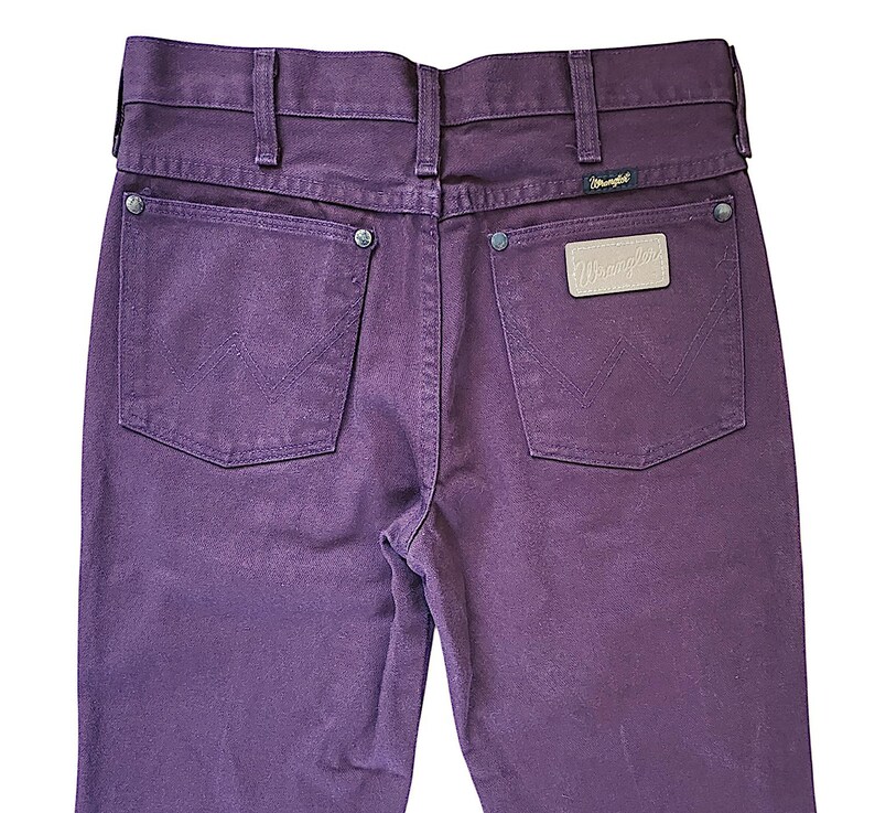 Puede incluir: Unos vaqueros de mezclilla de color morado intenso con cintura alta y trabillas para cintur&oacute;n, dos bolsillos traseros con remaches de metal plateado y un dise&ntilde;o de costura en W. Un parche de cuero beige con el logo Wrangler est&aacute; cosido en el bolsillo derecho.