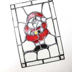 May include: A rectangular stained glass panel featuring a stylized Santa Claus figure. The design uses red, white, black, and yellow glass pieces held together by dark metal solder lines. A small metal loop is attached to the top for hanging.