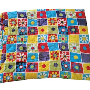 May include: A quilted blanket featuring a grid pattern of squares in bright colours including red, yellow, blue, and green. The squares are decorated with either a stylised flower graphic with white petals or a solid coloured circle in the centre.