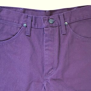 Puede incluir: Unos pantalones vaqueros de color morado intenso con dise&ntilde;o de cintura alta, cierre de botones, estilo de cinco bolsillos y remaches met&aacute;licos de color plateado en las esquinas de los bolsillos delanteros. La tela tiene una textura visible.