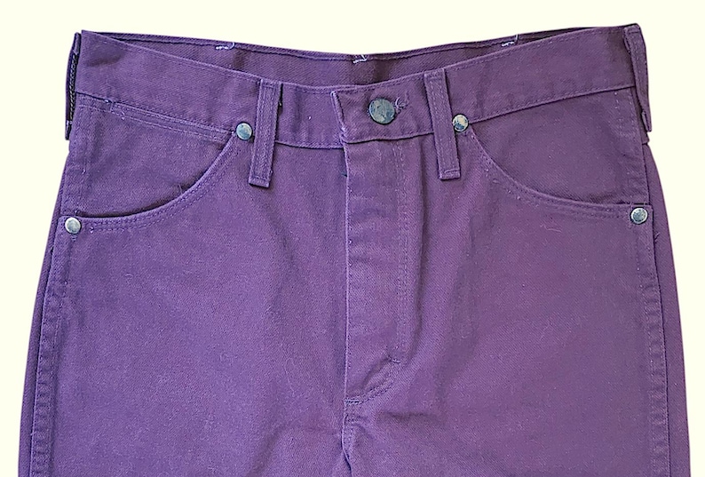 Puede incluir: Unos pantalones vaqueros de color morado intenso con dise&ntilde;o de cintura alta, cierre de botones, estilo de cinco bolsillos y remaches met&aacute;licos de color plateado en las esquinas de los bolsillos delanteros. La tela tiene una textura visible.