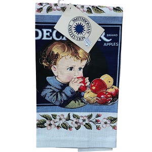 May include: A decorative kitchen towel featuring a vintage illustration of a child eating an apple, with a bowl of apples. The towel has a blue border with floral accents and the text "DEC BRAND APPLES". A Smithsonian Institution tag is attached.