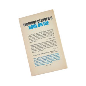 May include: The back cover of the book "Soul on Ice" by Eldridge Cleaver. The title is in blue, with the author's name above it. The cover has quotes and text in black. The book is on a white background.