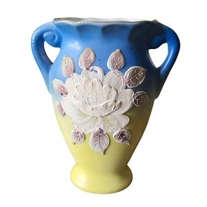 May include: A ceramic vase with a blue top and yellow base, featuring a white rose and leaf design. The vase has two handles and is approximately 20 cm tall. The floral design is raised and detailed.