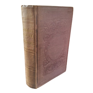 May include: A vintage hardback book titled Combe's System of Phrenology with a faded brown cloth cover. The spine features gold embossed lettering, and the front cover displays an intricate, debossed floral border pattern surrounding a blank centre.