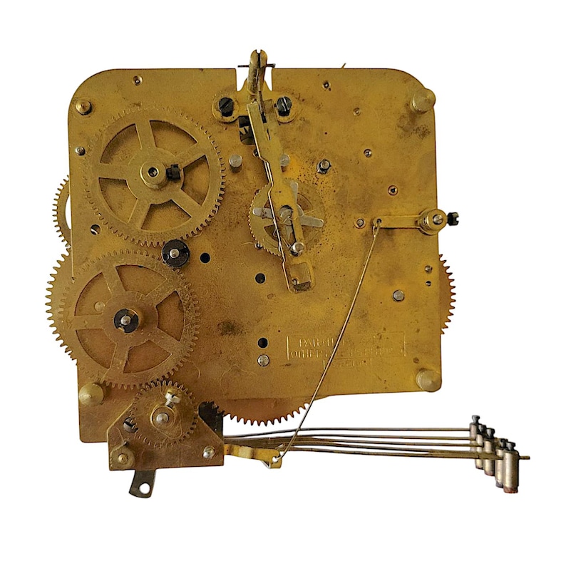 May include: Close-up of a vintage clock movement with brass gears and components. The mechanism features multiple interlocking gears, levers, and a pendulum weight system. The brass components have a warm, aged patina, suggesting the clock's age and history.