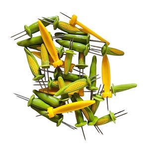 May include: A collection of corn-on-the-cob holders featuring green husks and yellow kernels. Each holder has two sharp metal prongs extending from the base, designed to secure corn while eating. The set includes multiple individual pieces.