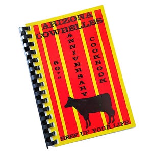 May include: A spiral-bound cookbook titled "Arizona Cowbelles 60th Anniversary Cookbook." The cover is red with yellow vertical stripes and features a black silhouette of a cow. The text "Beef Up Your Life" is at the bottom.