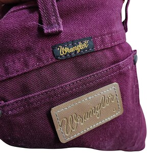 Puede incluir: Unos vaqueros de mezclilla de color morado intenso que presentan una etiqueta de tela negra con el logotipo amarillo de Wrangler cosido cerca de la cintura y un parche de cuero tostado con el nombre de la marca Wrangler bordado.