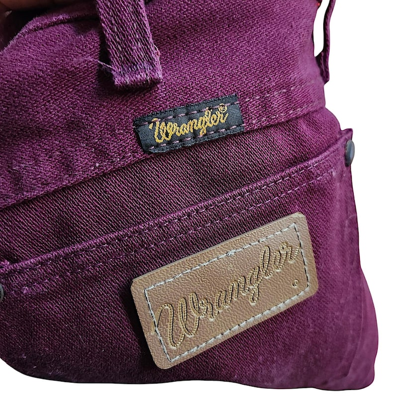 Puede incluir: Unos vaqueros de mezclilla de color morado intenso que presentan una etiqueta de tela negra con el logotipo amarillo de Wrangler cosido cerca de la cintura y un parche de cuero tostado con el nombre de la marca Wrangler bordado.